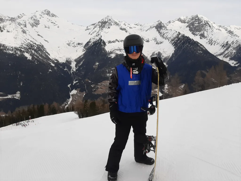 Ein Jugendlicher steht mit Snowboard auf der Piste, dahinter die Bergkette.